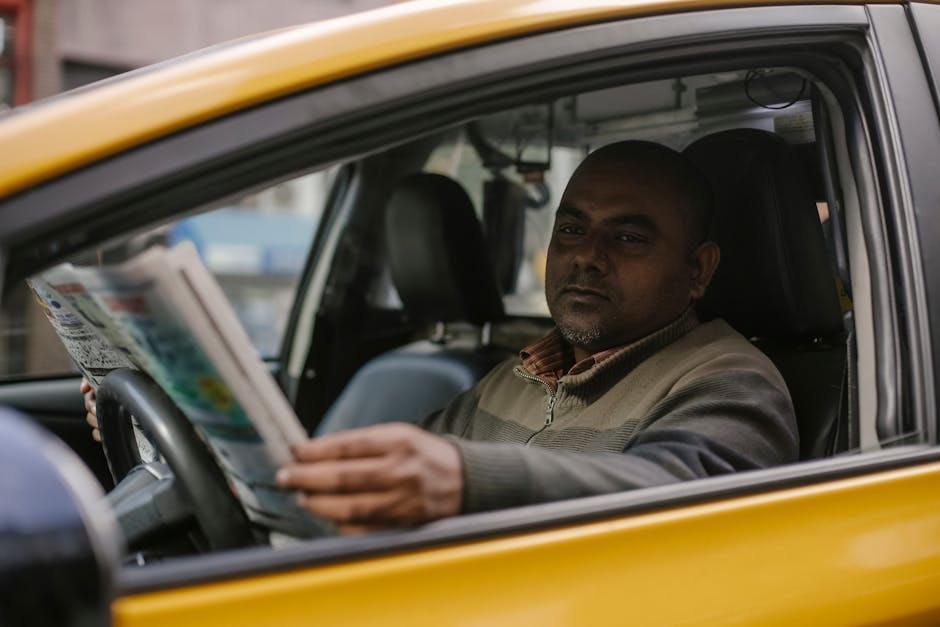 Concentrated ethnic male driver sitting at steering wheel and reading magazine while waiting for client